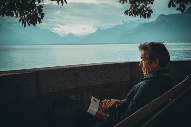Elderly man sitting on a bench by a lakeside, reading a book with mountains in the background.